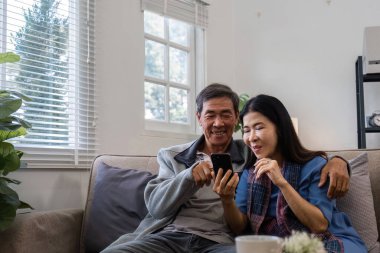 Emekli bir çift kanepede oturuyor, gülümsüyor ve akıllı telefona bakıyor, rahat bir oturma odasında birlikte vakit geçiriyorlar..