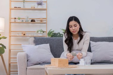 Genç bir kadın, rahat bir oturma odasında kanepede oturup hastalığını yatıştırmak için ilaç alıyor. Odaklanmış ve iyileşmeye kararlı görünüyor..