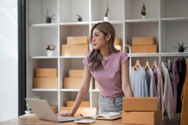 Genç bayan girişimci, çevrimiçi kıyafet işini envanterle çevrili şık bir ev ofisinden yönetiyor ve bir dizüstü bilgisayar üzerinde çalışıyor..