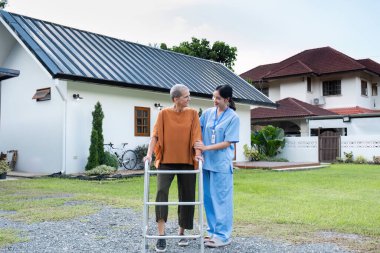 Yaşlı bir kadın ve bakıcısı evlerinin bahçesinde dolaşır, temiz havanın ve doğanın tadını çıkarır, yaşlı bakım ve desteği sembolize eder..