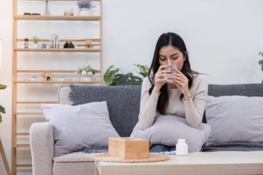 Kanepede oturan genç bir kadın, elinde bir bardak su ile ilaç alıyor ve kendini iyi hissetmediğini gösteriyor..