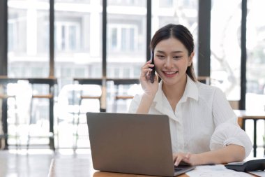 Kendine güvenen genç bir ofis çalışanı, modern bir ofiste dizüstü bilgisayarlı bir masada otururken telefonla iş hakkında konuşur. 