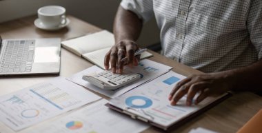 Evden çalışan bir işadamı, bilgisayarlı ve ahşap masadaki hesap makinesiyle finansal grafikleri ve belgeleri analiz ediyor..