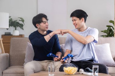 İki genç çocuk, rahat bir oturma odasında video oyunu oynarken, yumruk tokuştururken ve eğlenirken görülüyor..