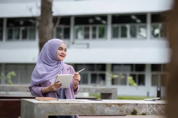 stock image A young Muslim student wearing a hijab, studying outdoors with a tablet at a university campus, symbolizing education, diversity, and modern learning.