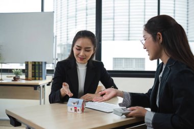 Modern bir ofiste iki emlakçı bir model evle emlak satışının detaylarını tartışıyor..