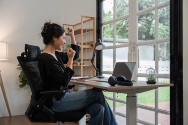 Geniş pencerelerden görülebilen doğal ışık ve yeşillik ile çevrili modern bir ev ofisinde finansal belgeler üzerinde gayretle çalışan genç bir muhasebeci..