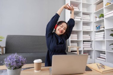 Rahat poz veren genç bir kadın, arka planda kahve ve modern ev dekoruyla birlikte kulaklık takarak sosyal medyayı keşfediyor..