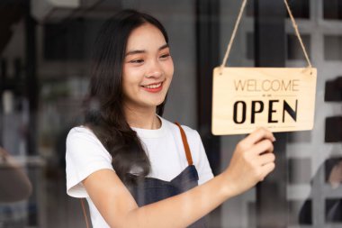 Önlüklü gülen kafe çalışanı modern bir kafenin girişine açık tabela asıyor. Müşterileri karşılamaya hazır..