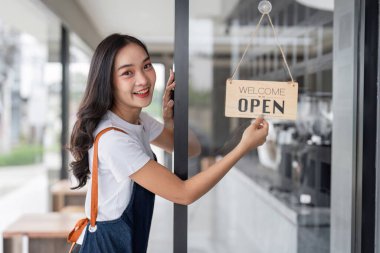 Neşeli genç bir barista modern bir kahve dükkanının girişinde duruyor. Gülümseyen ve açık bir tabelayla müşterileri karşılıyor..