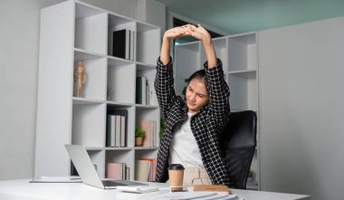 Genç bir iş kadını, modern bir ofis ortamında masasında otururken kendini işten yorgun hisseder..