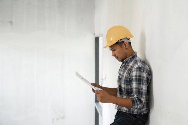 Genç bir mühendis güvenli bir şekilde çalışmasını planlıyor, elinde planlar tutuyor ve modern bir inşaat alanında güvenlik donanımı takıyor..