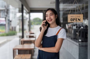 Güzel, genç bir barista modern bir kahve dükkanının girişinde duruyor. Gülümsüyor ve müşterileri karşılamaya hazır..