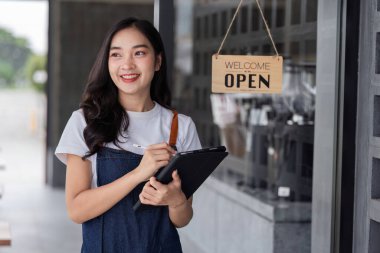 Modern bir kahve dükkanının girişinde duran genç ve güzel bir barista müşterileri sıcak bir gülümsemeyle karşılamaya hazır..