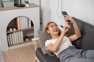 Genç bir kadın modern bir oturma odasındaki kanepeye uzanır, gülümser ve akıllı telefonunu kullanır, rahatlamanın tadını çıkarır..