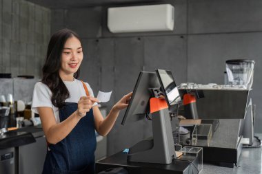 Modern bir kahve dükkanında çalışan genç bir barista yazar kasayı işletiyor ve müşteri memnuniyetini dostça bir tavırla sağlıyor..