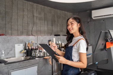 Modern bir kafede sipariş alırken gülümseyen genç bir barista. Önlük giyiyor ve modaya uygun bir kahve dükkanında çalışıyor..