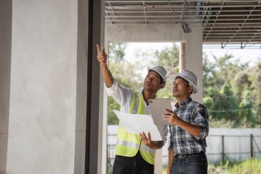 İnşaat çalışmalarını tartışan ve planlayan çelik yelek ve başlıklı mühendisler bir inşaat alanının planlarını ellerinde tutuyorlar..