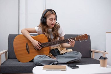 Şarkılar yazan, akustik gitar çalan, kulaklık takan ve çağdaş bir oturma odasında bir koltukta oturan yetenekli genç bir kadın sanatçı..