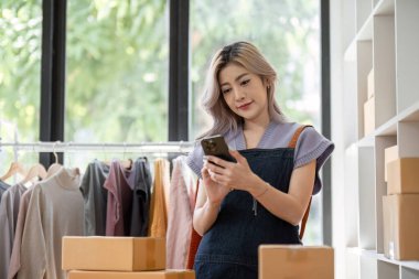 Genç bir bayan girişimci, modern bir ev ofisinde giysi rafları ve nakliye kutularıyla çevrili online gömlek satışlarını yönetmek için akıllı telefonunu kullanıyor..