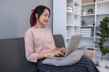 Genç bir kadın rahat bir şekilde kanepeye oturmuş, modern bir ev ortamında dizüstü bilgisayarında sosyal medyayı inceliyor..