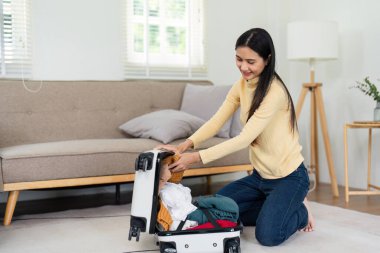 Bir kadın yere diz çöker, modern bir oturma odasında bavulunu toplar, seyahate ya da tatile hazırlanır. Gülümsüyor ve elbiselerini düzenliyor..