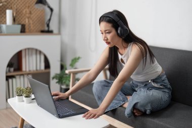 Genç bir kadın, modern bir oturma odasındaki kanepede rahat bir şekilde otururken, dizüstü bilgisayar ve kulaklık kullanarak evden ders çalışıyor..