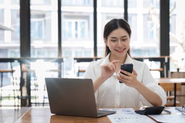 Modern bir ofiste genç bir kadın muhasebeci müşteriyle iletişim kurmak için akıllı telefon kullanıyor. Elinde dizüstü bilgisayar ve belgelerle bir masada oturuyor..