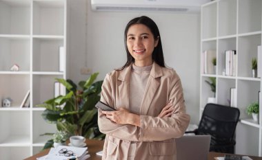 Kendine güvenen genç bir iş kadını modern bir ofiste duruyor, kollarını kavuşturup gülümsüyor, profesyonellik ve başarı yayıyor..
