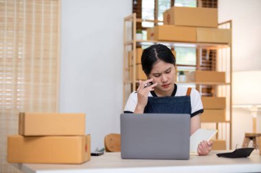 İnternette ürün satarken stresli hisseden genç bir kadın, evinde dizüstü bilgisayar ve paketleme kutularıyla çalışıyor..
