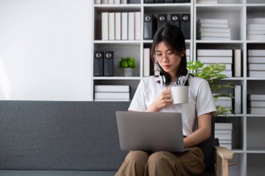 İnternette çalışmak için dizüstü bilgisayar kullanan, kulaklık takan ve elinde kahve fincanı olan genç bir kadın, modern bir evde bir kanepede oturuyor..