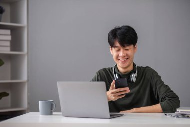 Evde online okuyan, dizüstü bilgisayar ve akıllı telefon kullanan, gülümseyen ve öğrenim ortamında meşgul bir çocuk..