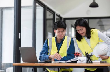 Güvenlik yelekli iki genç mühendis modern ofis ortamında bir masada inşaat projesi planlarını tartışıyor..