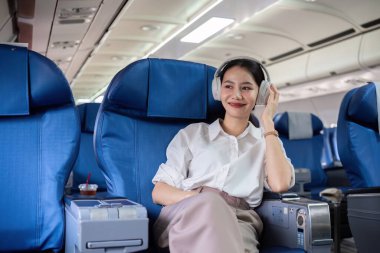 Genç bir iş kadını uçakta seyahat ederken kulaklıkla müzik dinliyor, rahat bir şekilde oturuyor ve gülümsüyor..