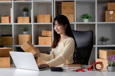 Bilgisayarında çalışan genç bir kadın online mağazasının müşteri siparişlerini kontrol ediyor. Etrafı nakliye kutuları ve ofis malzemeleriyle çevrili..