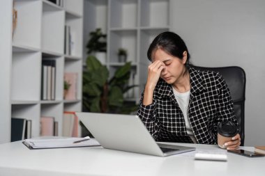 Bir iş kadını evde çalışırken kendini stresli ve bitkin hisseder, bir masada dizüstü bilgisayarla oturur ve elinde kahve fincanı taşır..