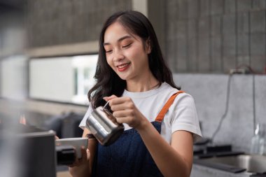 Modern bir kahve dükkanında kahve hazırlarken gülümseyen genç bir barista beyaz gömlek ve kot önlük giyiyor..