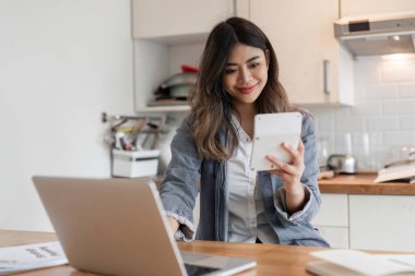 Modern bir mutfak ortamında dizüstü bilgisayar ve tablet kullanan evden çalışan genç bir serbest çalışan. İşine odaklanmış ve meşgul görünüyor..