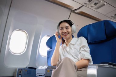 Genç bir iş kadını, uçakta seyahat ederken kulaklıkla müzik dinler, rahat bir koltukta gülümser ve dinlenir..