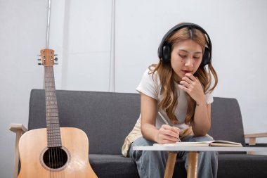 Genç bir kadın sanatçı kulaklık takıyor, yanında gitarla modern bir salonda şarkı yazıyor..