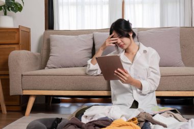 Genç bir kadın, bavulunu geziye hazırlarken gergin görünüyor. Giysileri etrafa saçılmış ve elinde bir tabletle yerde oturuyor..