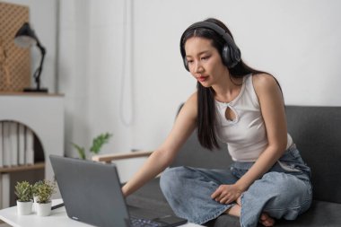 Genç bir kadın, modern bir oturma odasında rahat bir şekilde otururken dizüstü bilgisayar ve kulaklık kullanarak internet üzerinden ders çalışıyor..