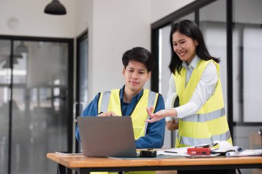 Güvenlik yelekli iki genç mühendis, bir dizüstü bilgisayar ve belge kullanarak modern bir ofis ortamında inşaat projesi planlarını tartışıyor..
