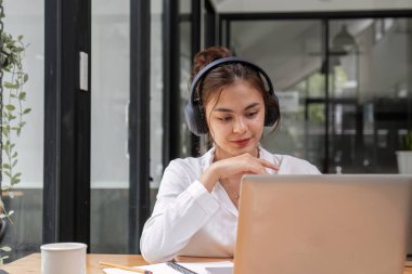 Genç bir iş kadını çevrimiçi bir toplantıya katılıyor, kulaklık takıyor ve çağdaş ofis ortamında dizüstü bilgisayarı üzerinde çalışıyor..