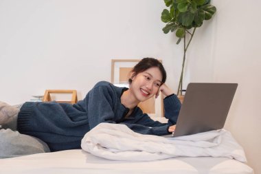 Genç bir kadın, minimalist dekoru ve yeşil bitkisi olan rahat, modern bir evde dizüstü bilgisayar kullanırken yatağında dinleniyor..