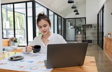 Genç bir kadın muhasebeci modern bir ofiste dizüstü bilgisayar kullanarak ve bir fincan kahvenin tadını çıkararak mutlu bir şekilde çalışıyor..