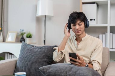 Genç bir adam modern odasında rahat bir şekilde oturup, kulaklıkla müzik dinlerken, dinleniyor ve telefonuyla oynuyor..