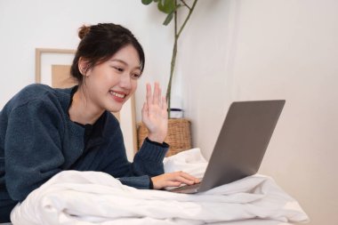 Evde dizüstü bilgisayar kullanan, gülümseyen ve el sallayan, rahat ve rahat bir yaşam tarzını, uzaktan çalışmayı ya da online iletişimi temsil eden genç bir kadın..