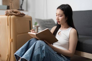 A young woman sits on the floor next to a suitcase, writing in a notebook as she prepares for her upcoming trip. clipart