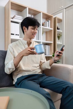 Genç bir adam odasında rahatça oturup, bir fincan kahvenin tadını çıkarırken telefonuyla oynuyor..